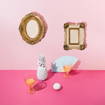 Tropical background with white pineapple and lemon, drinking glasses and blue paper fan on pink table and golden vintage picture frames on pink wall. Minimal concept.