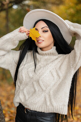 Happy beautiful young woman model with smile in white knitted sweater and hat holds a autumn yellow leaf and walks in fall colorful park