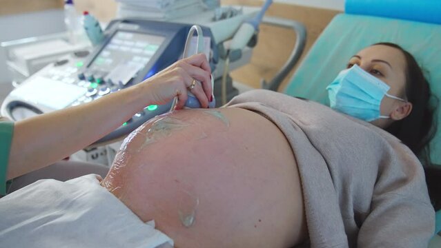 Pregnant woman in mask lies on the doctor's couch. Doctor moves the equipment around the belly with gel. Close up.