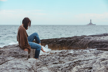 woman sitting by rocky sea beach in wet jeans lighthouse on background