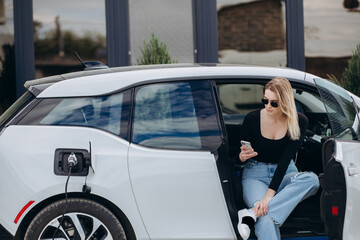 Charming woman sitting inside charging electric car with modern smartphone and cup of coffee in hands. Caucasian female talking by video call while waiting her auto to be charged.