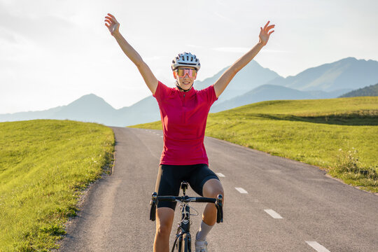 Female cycle race rider pedaling on an open road route in mountain landscape, passing by a car and raising arms in a victory pose, wide shot.