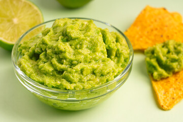 Close up view of mexican dip, spread or salad made of pureed ripe avocado served in glass bowl with crispy tortilla chips or nachos and halved lime slices as snack or appetizer on green background