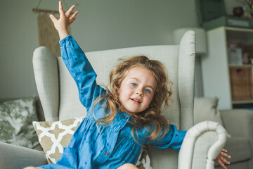 A little cute blue-eyed girl with curly long hair sits in a cozy armchair at home. Cozy, bright...