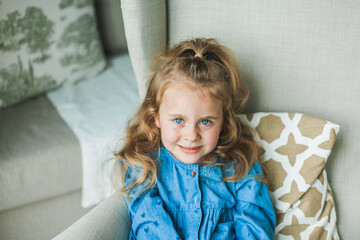 A little cute blue-eyed girl with curly long hair sits in a cozy armchair at home. Cozy, bright...
