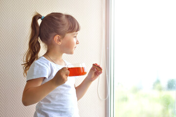 A girl in a white T-shirt holds a cup of tea and a liver in the form of a red heart standing by the window, watching. delicious sweet snack. safe at home, self-isolation during a pandemic.
