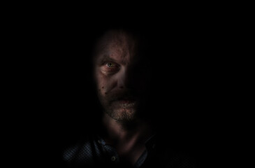 bald man with blond beard coming out of the darkness. The light shines on only half of the man's face and also illuminates the shirt neck. The man has three freckles on the illuminated part.