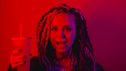 Young woman holds large cup of soft drink with straw in dark room. Portrait of female posing with nonalcoholic beverage in darkness illumination of red light.