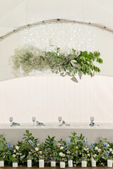 Wedding presidium in restaurant. Banquet table for newlyweds with flowers, greenery, candles and garland ligths. Lush floral arrangement. Luxury wedding decorations. Soft selective focus.