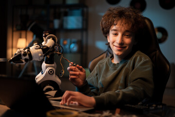 Portrait smiling boy spends evening time in room at desk, checks toy instructions on laptop, scrolls web pages, child pulls electronics cables from robot, fixes