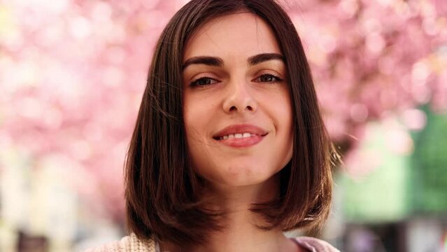 Portrait of a cute dark haired woman smiling charmingly while standing in the city street with sakura flowers in the background. Beautiful woman looking at camera outside.