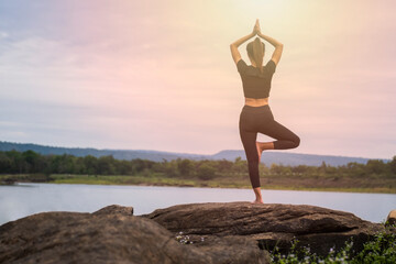 back of woman in gym clothes Doing yoga poses in tree pose on rocks with river and mountain views at sunset. concept of exercise in nature