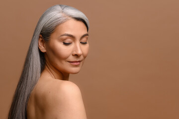 Headshot of attractive middle-aged Asian woman with naked shoulders and long silver hair isolated on brown, fascinating mature korean lady with smooth skin looks down dreamily. Skincare, copy space