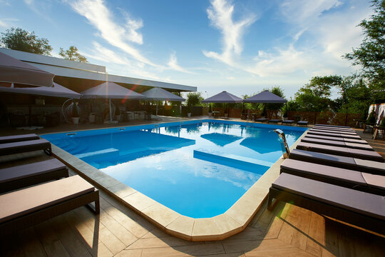 Swimming Pool Area With Deck Chairs And Sun Umbrellas Along The Poolside In Luxury Resort. Beach Beds Surrounded With Greenery And Blue Water. Tranquility And Relax Atmosphere On Vacation.