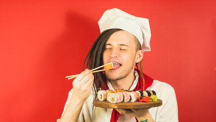 Young man dressed as chef holding wooden board with sushi rolls on red background. Male cook closed eyes eating appetizing sushi roll with chopsticks.