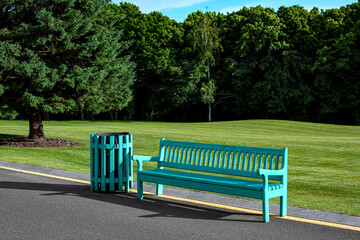 empty wooden bench with a trash can on an asphalt road with yellow markings and nature in the background city park, a hilly landscape with a green lawn and tall trees behind a clearing, nobody.