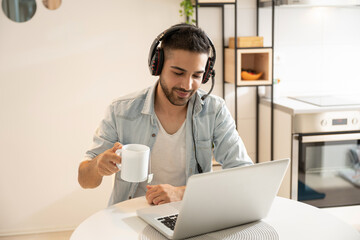 Portrait of bearded man wearing headphones with microphone drinking hot tea and using laptop at home.