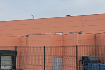 a large brown wall of an industrial building behind a green metal mesh fence against a gray sky...