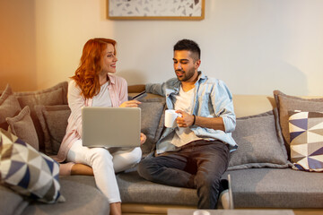 Young happy diverse loving couple relaxing on sofa and using laptop computer, holding credit card and shopping online at home. Beautiful redhead woman smiling and taking her boyfriend's credit card.