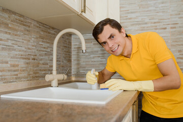 Young man in a yellow t-shirt and yellow gloves is cleaning the wash basin sink in the kitchen  using cleaning spray and sponge.