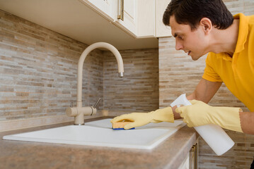 Young man in a yellow t-shirt and yellow gloves is cleaning the wash basin sink in the kitchen  using cleaning spray and sponge.