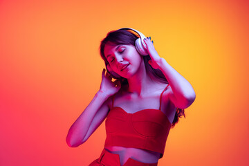 Portrait of young girl in headphones listening to music isolated over red-yellow background in neon...