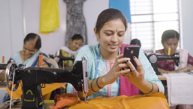 Woman tailor using mobile phone in front of sewing machine at garments - concept of relaxation, technology and checking online designs.