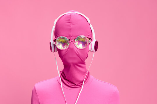 Art portrait of a woman wearing a pink full-face burglar mask with glowing round glasses wearing pink clothes with pink headphones on a pink background looking into the camera