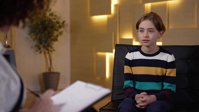 Contact preteen boy calmly answering questions of child counselor making notes during consultation. Cute little patient readily communicating with female therapist on appointment