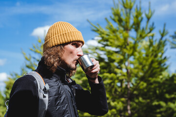 A tourist drinks coffee in the forest against the background of the blue sky, a serious guy drinks a glass of water looking ahead, a portrait of a young man on a journey through nature, a hipster