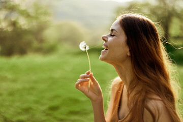 The woman smiles and blows the dandelion seeds into the wind. Summer and sunset light