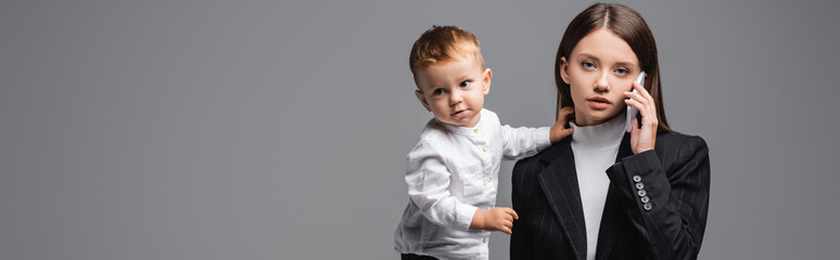 woman in black blazer talking on mobile phone near little son isolated on grey, banner.