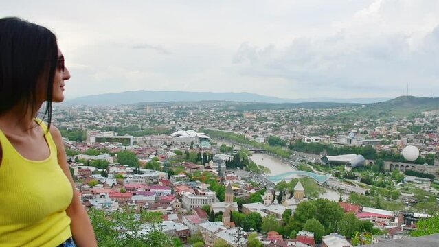 Panning view young caucasian woman stand on viewpoint enjoy scenic Tbilisi city panorama from Narikala fortress in summer. Travel in Georgia, caucasus