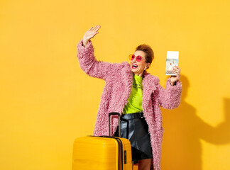 Happy woman is ready to travel. Photo of elderly woman wears pink glasses, dressed in fashionable outfit, with tickets and suitcase on yellow background