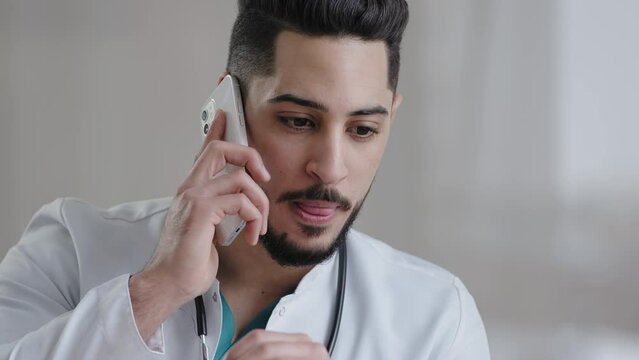 Serious male hispanic guy doctor therapist medical assistant consult patient distant discuss issues by mobile phone arab man adviser surgeon provide remote consultation use cellphone at clinic cabinet