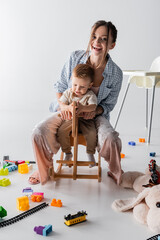 excited woman looking at camera while riding rocking horse with son on white.