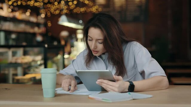 Beautiful caucasian brunette looking through some papers while sitting tafe with digital tablet hands. Young woman in casual attire spending leisure to for studying.