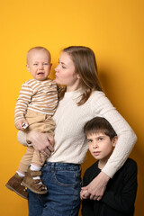 A mother with two sons on a yellow background. A crying baby and a cranky boy hug his mother.