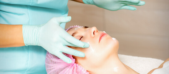 Fototapeta premium Cosmetologist with gloved hands applies a moisturizing mask with peeling cream on the female face. Facial cosmetology treatment. Procedures for facial care