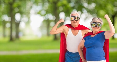 Portrait of caucasian senior couple wearing red capes and masks flexing muscle against trees in park - Powered by Adobe