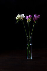 A bouquet of freesia stands in a transparent glass vase on a wooden table. Black background, low key, vertical orientation.
