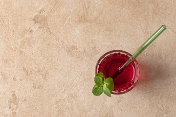 Top view of karkade tea (hibiscus) poured into a glass with a green glass tube and mint leaves. Horizontal orientation, copy space, beige textured background.