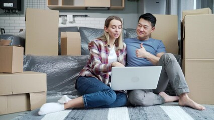 Young family in a new apartment after moving among the cardboard boxes sitting on the floor and happily choosing new furniture online store, use laptop for shopping. after relocation in home, house