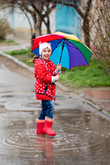 A cute little girl in a red cape, red boots and a white hat jump in puddles and has a fun.The girl has a rainbow umbrella in her hands. Happy childhood. Early spring. Emotions.