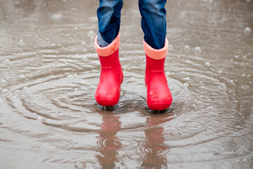 A cute little girl red boots jump in puddles and has a fun. Happy childhood. Early spring. Emotions.