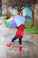 A cute little girl in a red cape, red boots and a white hat jump in puddles and has a fun.The girl has a rainbow umbrella in her hands. Happy childhood. Early spring. Emotions.