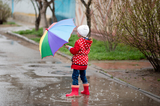 A cute little girl in a red cape, red boots and a white hat jump in puddles and has a fun.The girl has a rainbow umbrella in her hands. Happy childhood. Early spring. Emotions.