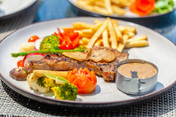 Pork chop serve with vegetables, french fries and pepper sauce. Grilled pork steak with caramelized pear on white plate in outdoor restaurant. Gourmet food