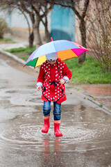 Obraz premium A cute little girl in a red cape, red boots and a white hat jump in puddles and has a fun.The girl has a rainbow umbrella in her hands. Happy childhood. Early spring. Emotions.