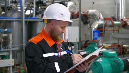 A male engineer in a white helmet at the workplace holds a folder with a blueprint of the technological process of the production line of a gas compression plant in his hands.